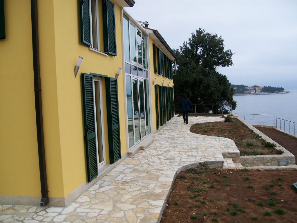 Osoba hoda kamenim putem uz žutu zgradu sa zelenim kapcima, smještenu pokraj vodene površine i drveta.