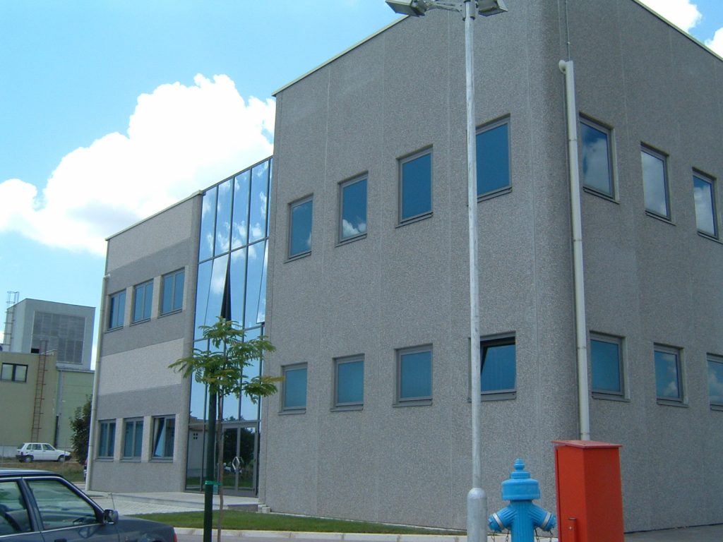 Moderna siva dvoetažna uredska zgrada s više kvadratnih prozora i staklenim ulaznim dijelom; u prvom planu nalaze se plavi hidrant i crvena komunalna kutija.