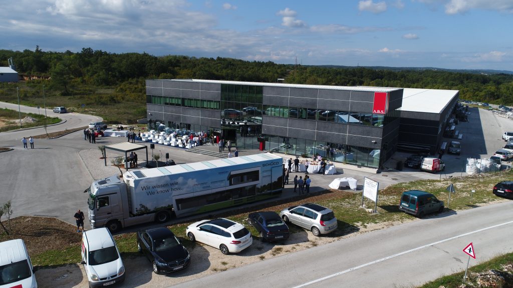 Moderna dvoetažna poslovna zgrada s velikim prozorima, ljudima okupljenima ispred, bijelim automobilima na parkiralištu i brendiranim kamionom ispred, pod djelomično oblačnim nebom.