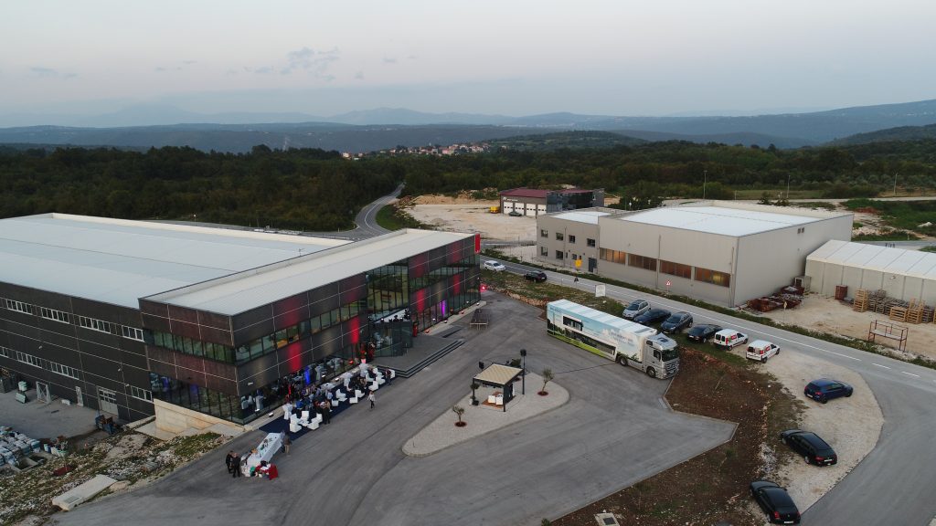 Zračni prikaz industrijskog kompleksa s dvije velike zgrade, nekoliko parkiranih automobila, autobusom i ljudima okupljenima ispred jedne zgrade; okružen zelenilom i brežuljcima.