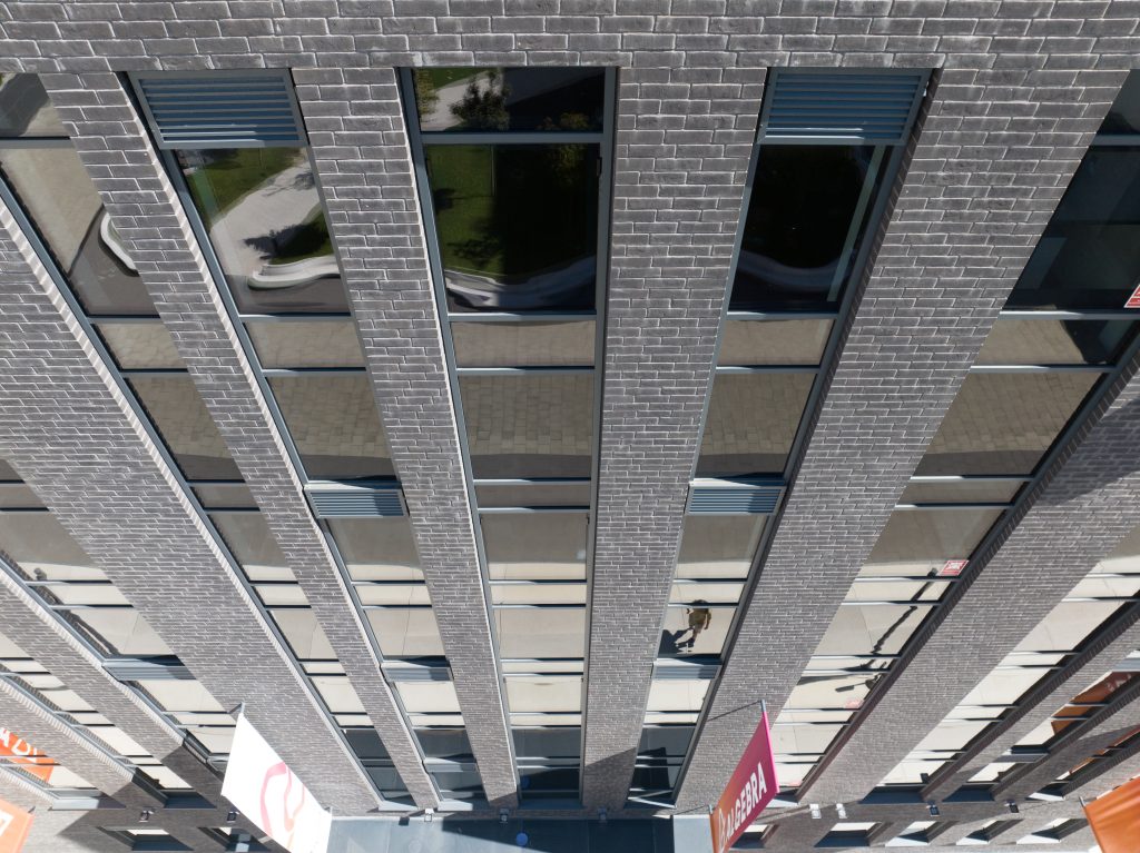 Tall brick building with large vertical windows reflecting the surroundings, seen from an overhead perspective. Banners are hanging at the entrance below.