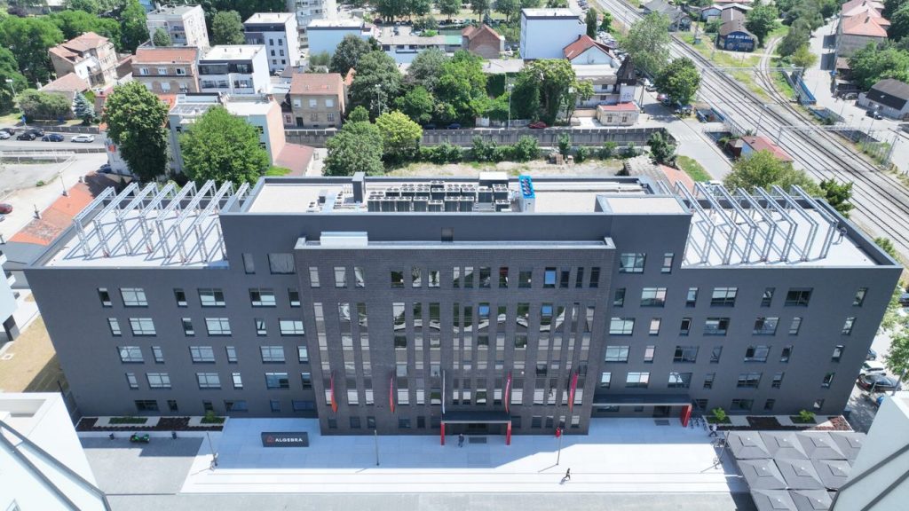 Zračni prikaz moderne, pravokutne sive zgrade s velikim prozorima i solarnim panelima na krovu, okružene drvećem i obližnjim stambenim objektima.