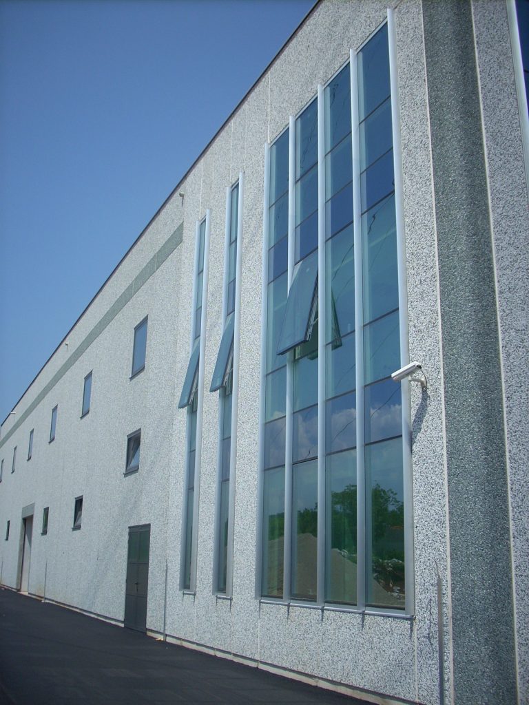 Velika industrijska zgrada s visokim okomitim prozorima, od kojih su neki otvoreni, i sivim teksturiranim vanjskim zidom pod vedrim plavim nebom.