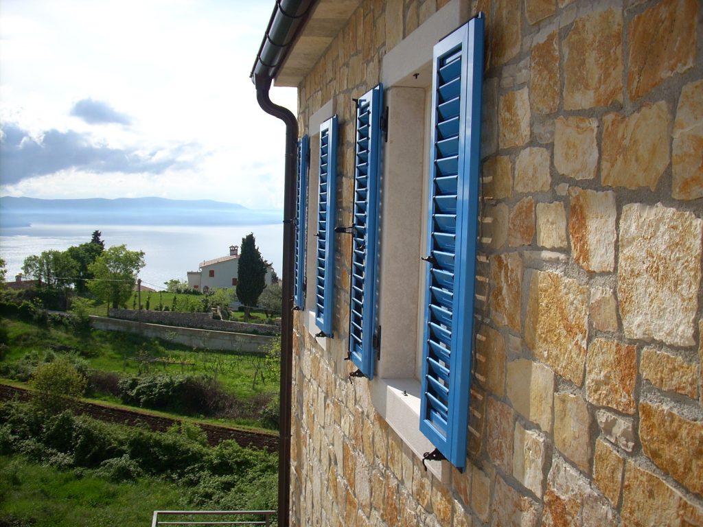 Kamena zgrada s plavim kapcima na prozorima gleda na travnati krajolik i vodenu površinu pod djelomično oblačnim nebom.