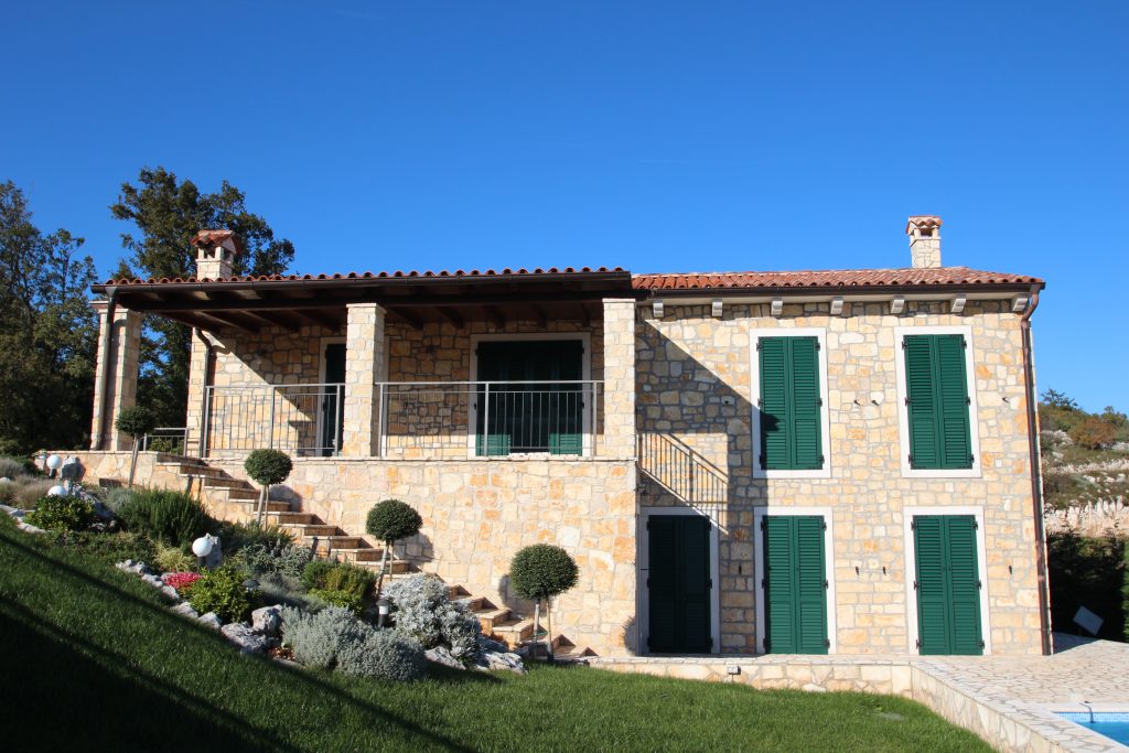 Stone house with green shutters, a tiled roof, and a front porch, surrounded by a landscaped garden on a sunny day.