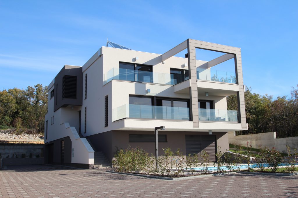 Moderna trospratna kuća s velikim staklenim balkonima, ravnim krovom i vanjskim stubištem, smještena uz popločani prilaz i uređen vrt.