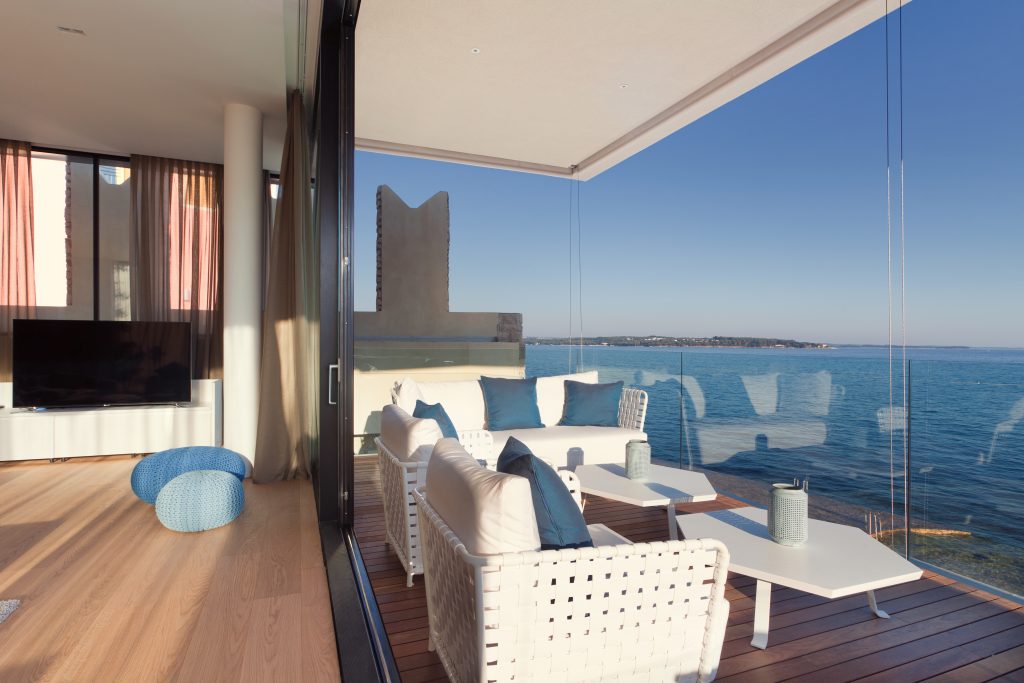 Moderan dnevni boravak koji se otvara na balkon sa staklenim zidovima, s bijelim namještajem od pruća i pogledom na ocean pod vedrim nebom.
