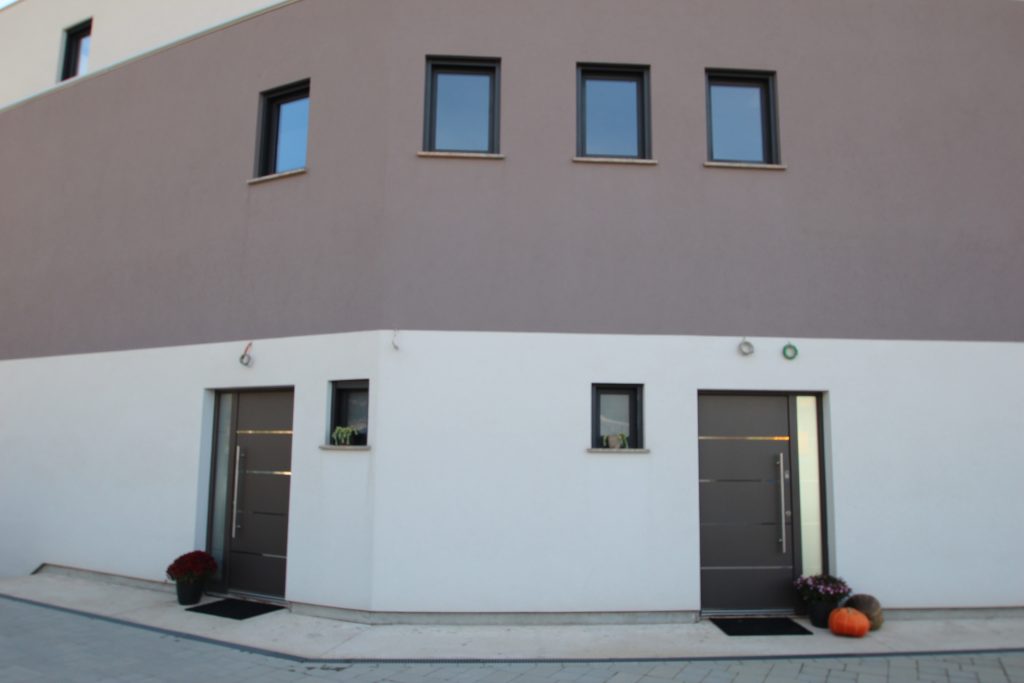 Vanjski dio moderne zgrade s dvama sivim vratima, tri mala gornja prozora i dva donja prozora. Pored vrata na popločanom putu nalaze se cvjetne posude i bundeva.