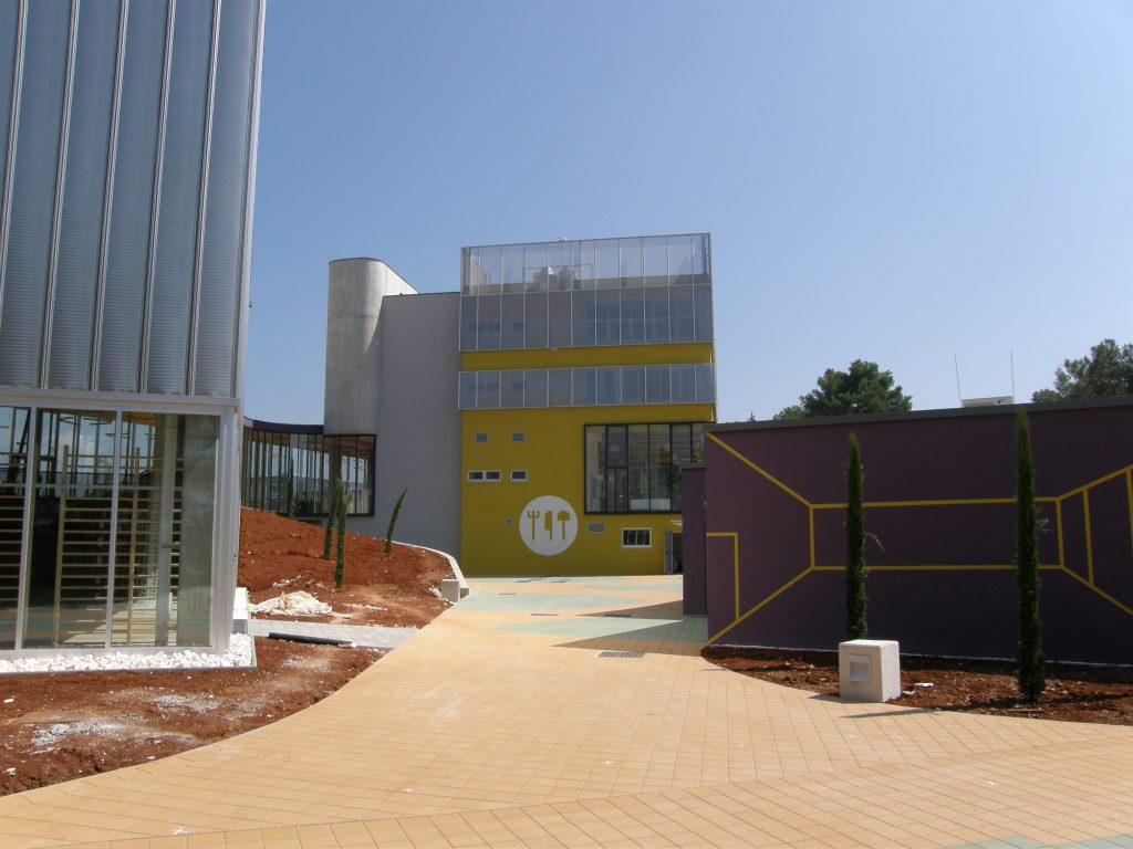 Moderna zgrada sa žutim pročeljem i staklenim panelima, okružena popločanim stazama i uređenim okolišem s crvenkastim tlom i mladim stablima, pod vedrim nebom.
