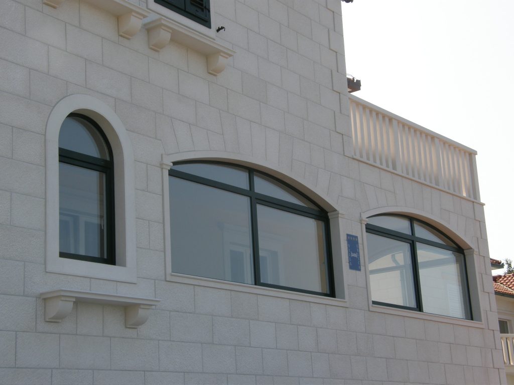 Blizak prikaz bijele kamene fasade zgrade s tri lučna prozora tamnih okvira i djelomičnim pogledom na balkon s bijelom ogradom.