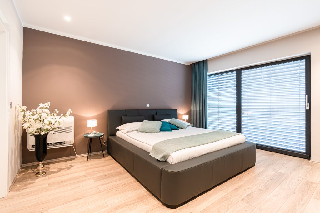 Moderna spavaća soba s velikim krevetom, dva noćna ormarića s lampama, vazom s cvijećem, drvenim podom i prozorima od poda do stropa s roletama.