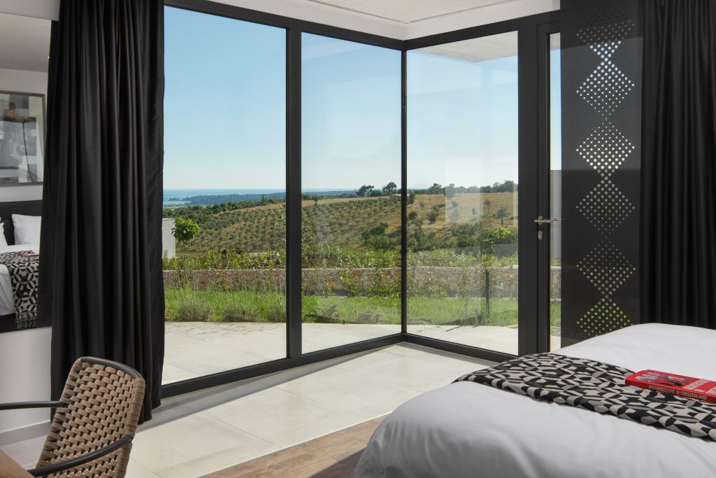 A modern bedroom with large glass doors overlooks a scenic landscape of fields and distant water under a clear sky.