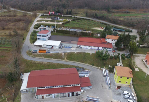Zračni prikaz manjeg industrijskog kompleksa s nekoliko zgrada, zakrivljenom cestom, raspršenim vozilima i okolnim zelenilom.