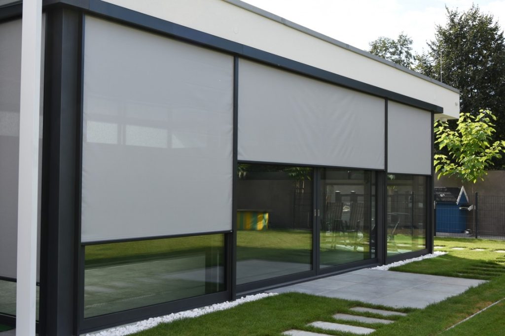 Modern building with large glass windows partially covered by gray exterior roller blinds, surrounded by a small grass lawn and stone pathway.