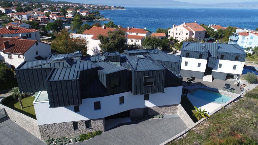 Zračni prikaz modernih stambenih zgrada s tamnim krovovima u blizini obale, okruženih kućama, zelenilom i bazenom.