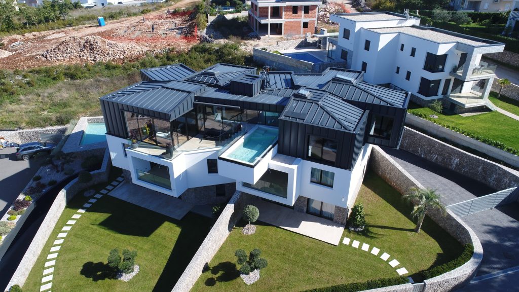 A modern multi-level house with angular architecture, large windows, and a rooftop pool is surrounded by landscaped lawns and situated on a sloped property.