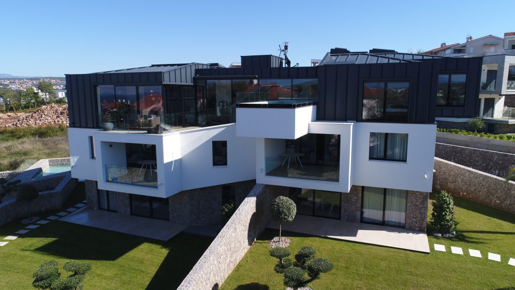 Modern multi-level house with large glass windows, white and dark exterior, surrounded by landscaped lawn and stone walls under clear blue sky.