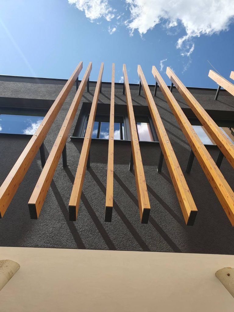 Drvene grede protežu se okomito s fasade moderne zgrade, a u pozadini se vide prozor i plavo nebo s oblacima.
