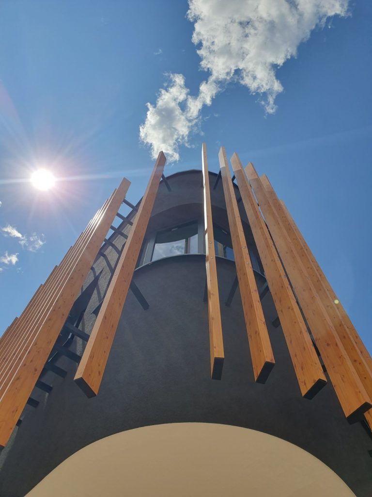 Fasada moderne zgrade s vertikalnim drvenim letvicama, zakrivljenim crnim zidom i okruglim prozorom, fotografirana odozdo prema plavom nebu sa suncem i oblacima.