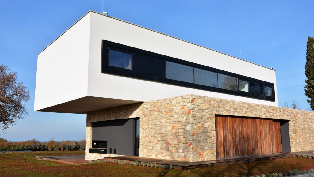 Moderna dvoetažna kuća s donjom etažom od kamena i drva te gornjom bijelom etažom koja ima dugi, uski prozor s crnim okvirom, smještena pod vedrim nebom.