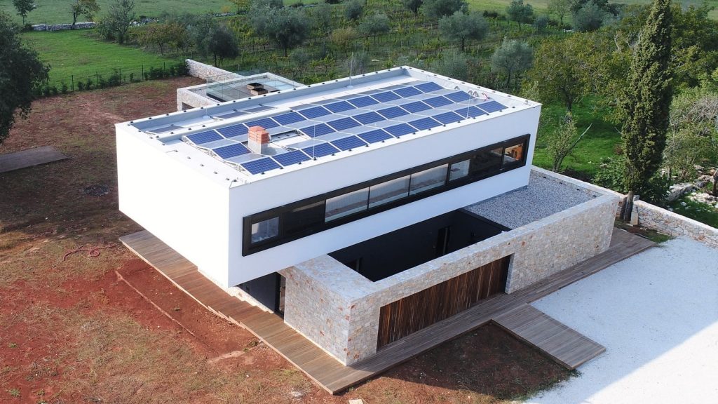 Moderna dvoetažna kuća sa solarnim panelima na ravnom krovu, okružena drvećem i crvenkastim tlom, s vanjskim elementima od kamena i drva.