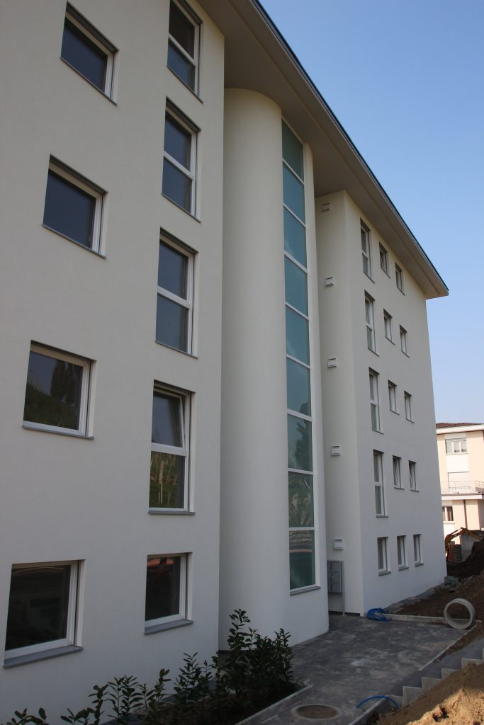 A modern four-story apartment building with white exterior walls, multiple windows, and a vertical glass section in the center.