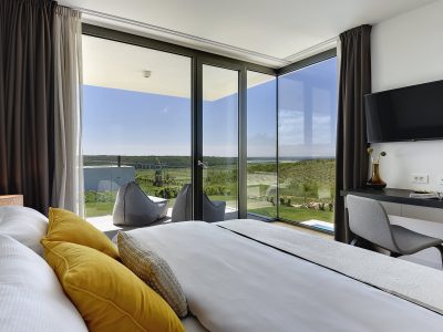 Moderna hotelska soba s uredno namještenim krevetom, radnim stolom i stolicom, televizorom na zidu te velikim prozorima s pogledom na travnati krajolik i plavo nebo.