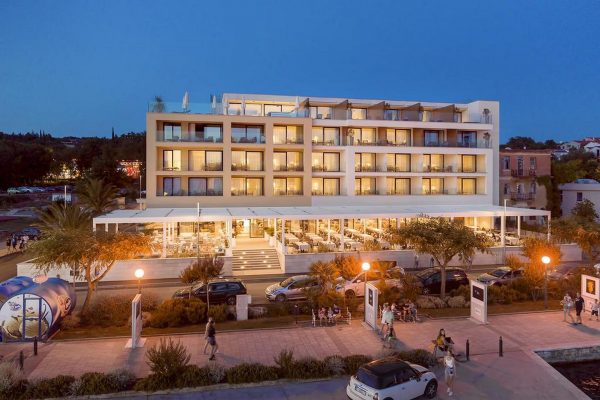 Moderan hotel na četiri kata s velikim prozorima i balkonima, vanjskim prostorom za objedovanje, parkiranim automobilima i prolaznicima koji hodaju nogostupom u sumrak.