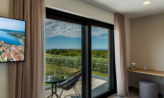 Moderna hotelska soba s balkonom na kojem se nalaze stol i stolice te pogledom na zelenilo i udaljeno more kroz velika staklena vrata. U sobi su vidljivi televizor i mali radni stol.