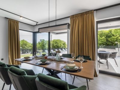 Moderna blagovaonica s drvenim stolom za šest osoba, zelenim baršunastim stolicama, velikim prozorima, zlatnim zavjesama i pogledom na terasu i zelenilo izvana.