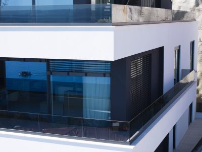 Moderna bijela zgrada s velikim staklenim prozorima plavkastog tona i crnim ogradama, koja reflektira vodu i vanjsko okruženje.
