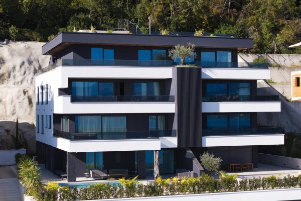 Moderna trospratna zgrada s velikim plavkastim prozorima, bijelom i tamnom vanjštinom, balkonima i biljkama na krovu, smještena uz padinu sa zelenilom.