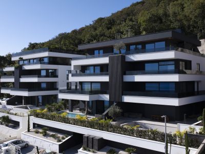 Moderne višekatne stambene zgrade s velikim prozorima i balkonima, izgrađene na padini s drvećem u pozadini.