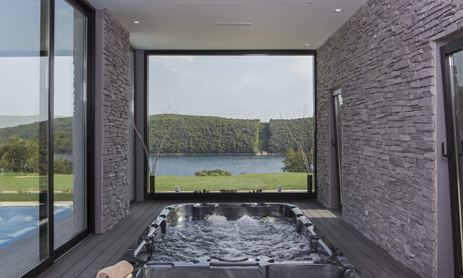 Unutarnji jacuzzi s mjehurićastom vodom, okružen kamenim zidovima i velikim prozorima koji gledaju na jezero i zelene brežuljke.