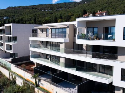 Moderne višekatne stambene zgrade s velikim staklenim balkonima, smještene na padini s zelenilom u pozadini pod vedrim nebom.