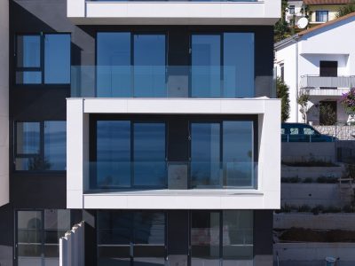 Moderna stambena zgrada s velikim staklenim balkonima i prozorima koji reflektiraju nebo; u pozadini su vidljive stambene kuće i stepenice.