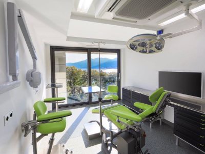 Moderna medicinska ordinacija s zelenim stolicama, medicinskom opremom, velikim monitorom i prozorom s pogledom na slikoviti vanjski prostor.