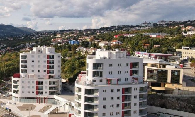 Dvije moderne bijele stambene zgrade s crvenim detaljima stoje uz cestu, okružene manjim zgradama i zelenim brežuljcima pod djelomično oblačnim nebom.