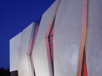 Moderna zgrada s kutnim, geometrijskim betonskim panelima i prozorima s crvenkastim odsjajem, fotografirana u sumrak na pozadini tamnoplavog neba.