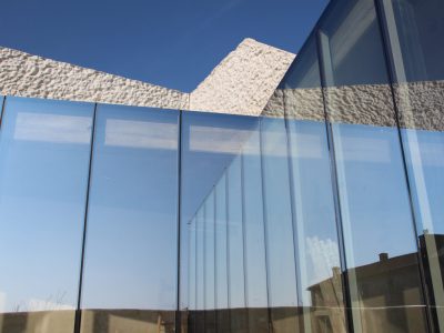 Moderna zgrada s visokim staklenim panelima koji reflektiraju nebo i teksturiranim bijelim betonskim elementima pod vedrim dnevnim svjetlom.