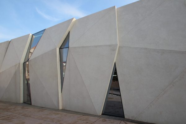 Fasada moderne zgrade s velikim geometrijskim betonskim panelima i uskim vertikalnim prozorima, s kutnim linijama i oštrim rubovima pod plavim nebom.
