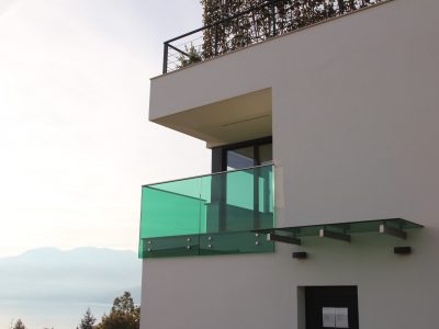 Moderna zgrada s balkonom od stakla i staklenim nadstrešnicom iznad vrata, s minimalističkim dizajnom i pogledom na udaljenu planinu i jezero.