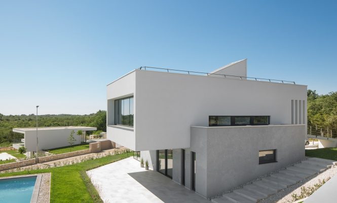 Modern two-story house with large windows, flat roof, and minimalist design; pool in the foreground and another similar building in the background under a clear blue sky.