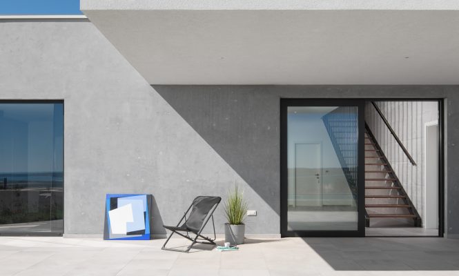 Moderan patio sa sivom betonskom vanjštinom, staklenim vratima s crnim okvirom, sklopivom stolicom, biljkom u posudi i uokvirenom umjetninom naslonjenom na zid.