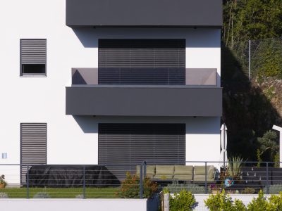 Moderna bijela zgrada sa zatvorenim sivim kapcima, balkonom, vanjskim prostorom za sjedenje, biljkama u teglama i prekrivenim predmetom u prvom planu.