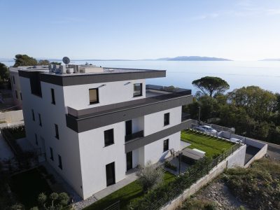 Moderna trospratna bijela stambena zgrada s ravnim krovom, balkonima i vrtnim prostorom, smještena u blizini velike vodene površine, s vidljivim udaljenim planinama i drvećem.