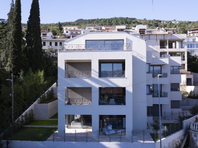 Moderna trospratna stambena zgrada s balkonima i velikim prozorima, okružena ogradom i uređenim okolišem, s drugim zgradama i drvećem u pozadini.