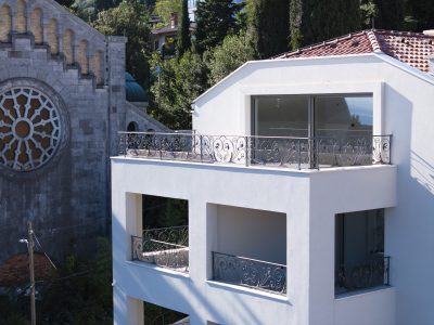 Moderna bijela zgrada s metalnim ogradama stoji pokraj starije kamene građevine s velikim kružnim prozorom od vitraja. U pozadini su vidljiva stabla i zelenilo.