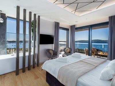 Moderna hotelska soba s velikim krevetom, dvjema stolicama, samostojećom kadom, prozorima od poda do stropa i pogledom na jezero ili more s brodovima i udaljenim brežuljcima.