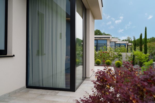 Modern house exterior with large glass windows, beige walls, and a patio surrounded by landscaped greenery and shrubs under a clear sky.
